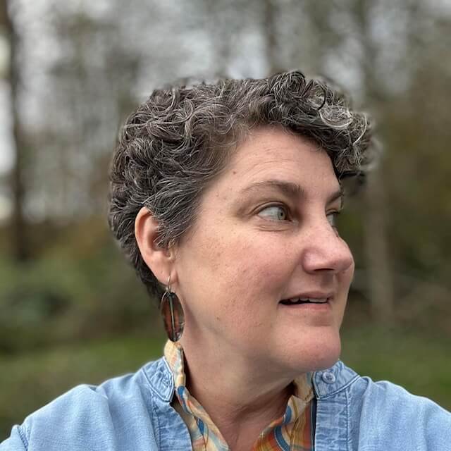 Kel Dylla, Rhapsody Project Council Member, with short curly brown and gray hair and a light blue denim shirt, looking off camera to the right, with trees in the background