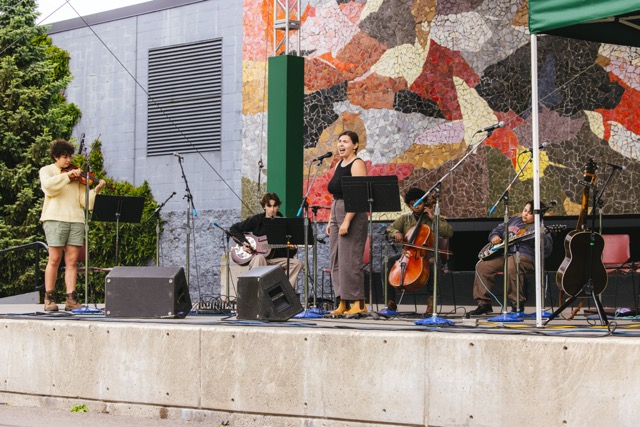 5 Songsters perform on the Mural stage at Seattle Center: Violin, resonator guitar, singer, cello, electric guitar.