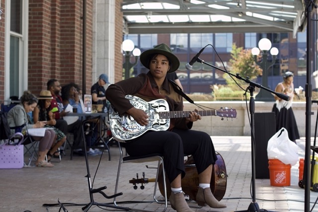Mariah seated in a chair on the King Street Station Plaza, playing a silver resonator guitar